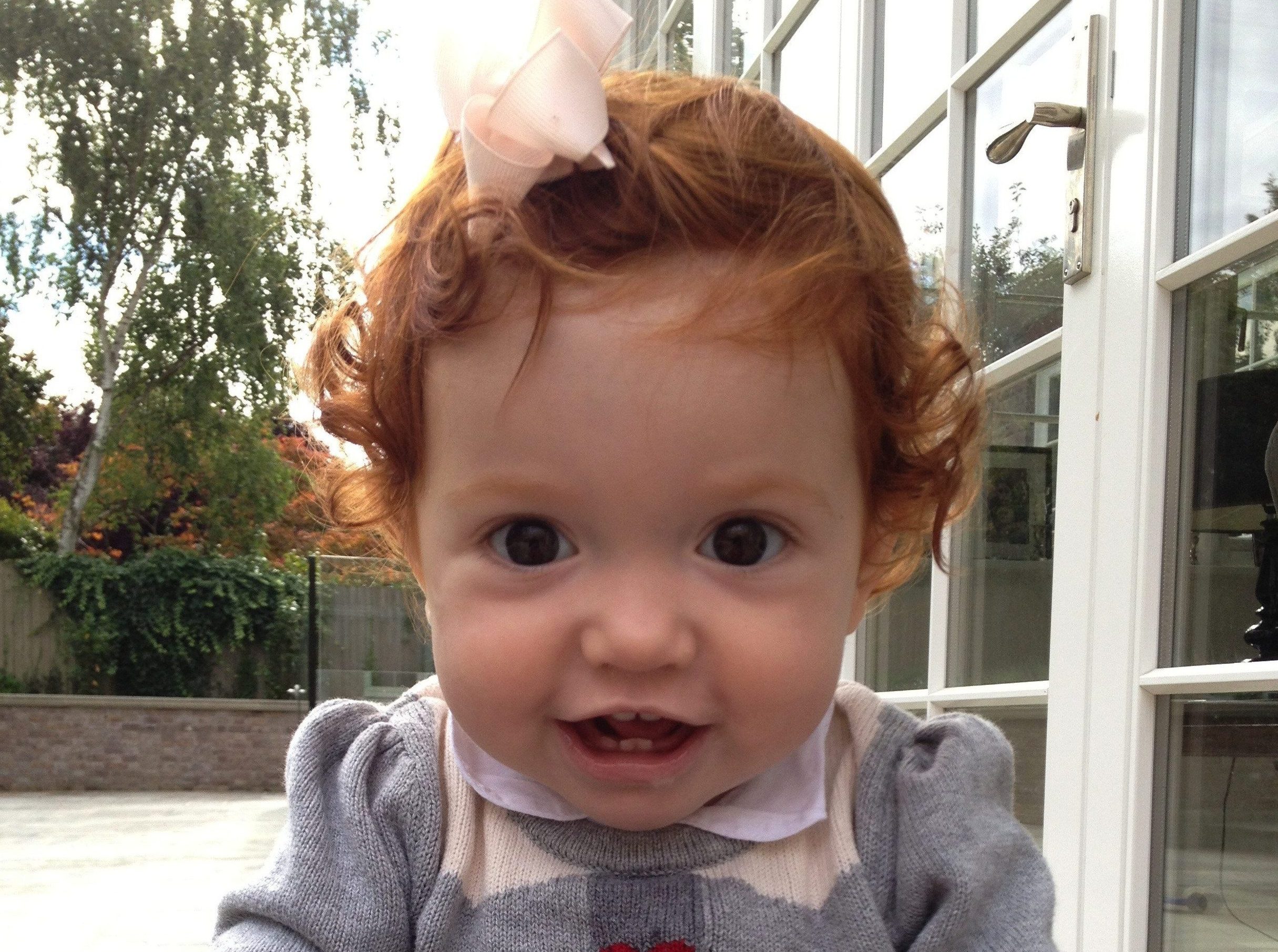 A joyful close-up of young Margot Martini, red curls and a pale pink bow, smiling outdoors.